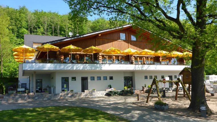 Ein Freizeitbereich mit einem großen Holzgebäude und sonnigen Terrassen. Gelbe Sonnenschirme bieten Schutz vor der Sonne, umgeben von Bäumen und einer freundlichen Umgebung.