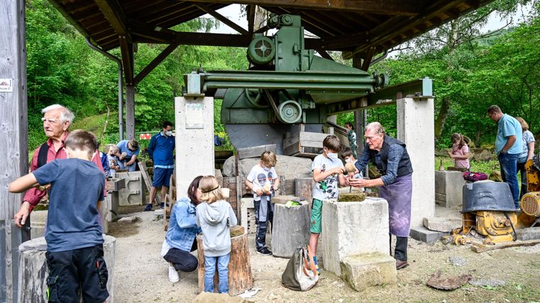 Eine Gruppe von Menschen, darunter viele Kinder, arbeitet unter einem überdachten Bereich an einer Steinbearbeitung. Im Hintergrund ist eine alte Maschine zu sehen, die für die Verarbeitung von Materialien verwendet wird.
