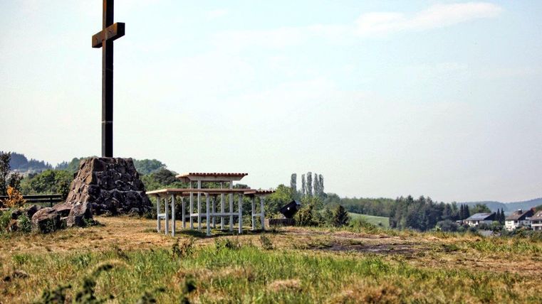 Ein großes Kreuz steht auf einem Hügel mit Blick auf die Landschaft. Daneben befindet sich eine kleine Sitzgruppe.