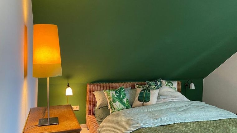 Ein gemütliches Schlafzimmer mit grün gestrichenen Wänden und einem modernen Bett. Die Beleuchtung wird durch eine Stehlampe und Wandlampen akzentuiert.
