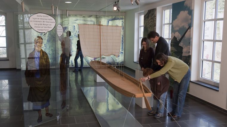 Eine Gruppe von Menschen steht in einem Ausstellungsraum und arbeitet an einem Modell eines Bootes. Im Hintergrund ist eine Wand mit Bildern und Texten zu sehen.