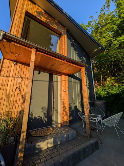 Ein modernes Haus mit Holzverkleidung und großen Fenstern. Vor dem Eingang stehen zwei weiße Stühle und ein kleiner Tisch.
