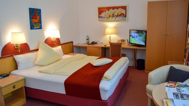 Ein gemütliches Hotelzimmer mit einem Doppelbett 180x200cm für 1 Person / Einzelnutzung. Es gibt einen Schreibtisch, einen Fernseher und einen kleinen Sitzbereich.