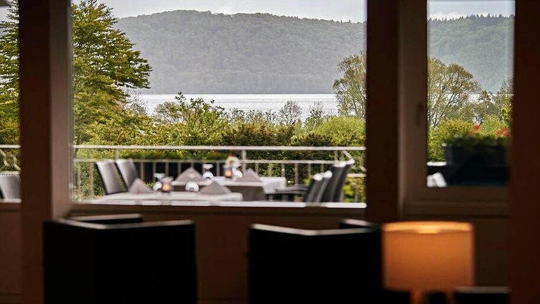 A view through a window onto an idyllic landscape with green spaces and a body of water in the background. In the foreground, there is table decor and chairs, indicating a cozy atmosphere.