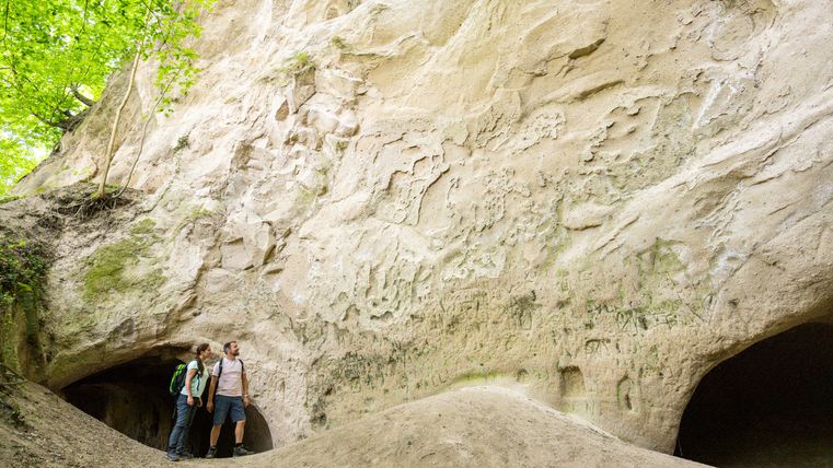 Zwei Personen stehen vor großen Felsen mit Höhlenöffnungen im Wald.