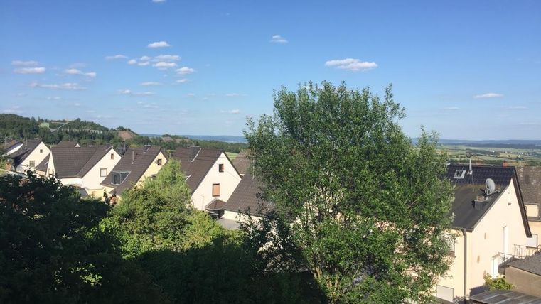 Eine ruhige Wohngegend mit modernen Häusern und einem großen Baum im Vordergrund. Im Hintergrund zieht sich eine sanfte Landschaft unter blauem Himmel.