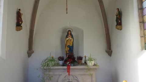 Eine kleine Kapelle mit einer Statue der Jungfrau Maria in der Mitte. An den Wänden sind zwei Heiligenfiguren und es gibt Pflanzen auf dem Altar.