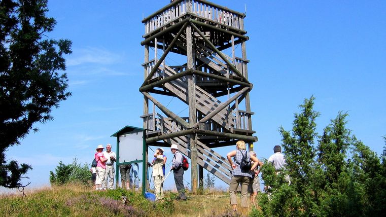 Ein Aussichtsturm steht in einer grünen Landschaft. Wanderer erkunden die Umgebung und genießen die Natur.