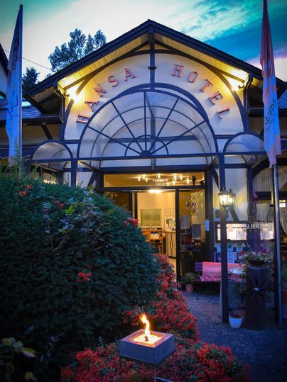 Ein einladendes Hotel mit dem Namen "Hansa" und einer stilvollen Fassade. Die Atmosphäre wird durch das sanfte Licht einer originellen Basaltkerze und die dekorativen Pflanzen im Vordergrund verstärkt.