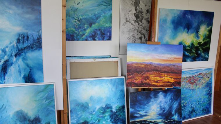 Eine Sammlung von verschiedenen Gemälden in Blau- und Grüntönen hängt an der Wand. Einige Bilder zeigen abstrakte Landschaften, andere Wasser- und Luftmotive.