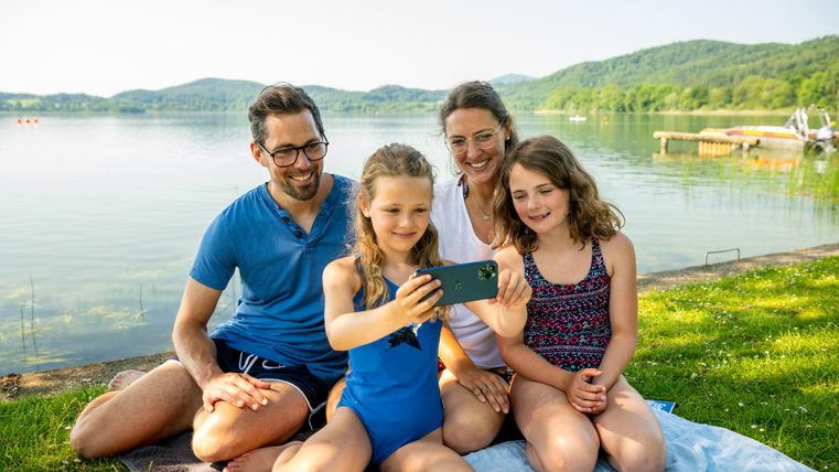 Eine Familie sitzt am Ufer eines Sees und schaut gemeinsam auf ein Smartphone. Die Kinder lächeln und genießen die Zeit miteinander in der Natur.