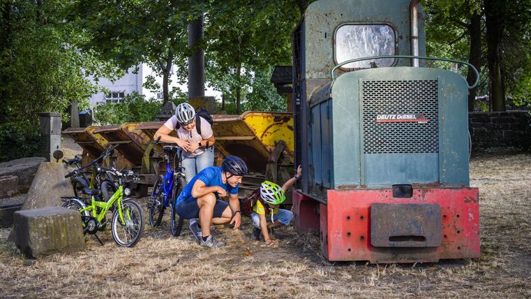A few children and adults are exploring an old locomotive. Bicycles are standing around them on dry, green ground.