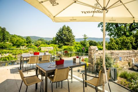 Ein einladender Außenbereich eines Restaurants mit Tisch und Stühlen. Im Hintergrund sind grüne Bäume und Hügel zu sehen, die eine entspannte Atmosphäre schaffen.