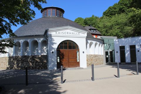 Ein historisches Gebäude namens Kaiserhalle mit einem runden Dach. Es liegt in einer grünen Umgebung und hat eine elegante Fassade.