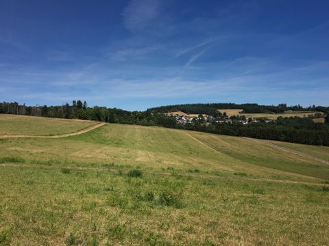 Een schilderachtig landschap met groene weilanden en een klein dorp op de achtergrond. Heuvels en bomen omringen de scène onder een bewolkte lucht.