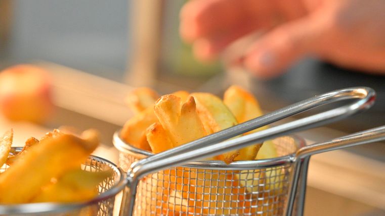 Eine Hand greift nach knusprigen Pommes frites in einem Frittierkorb. Die Pommes sind goldbraun und frisch zubereitet.