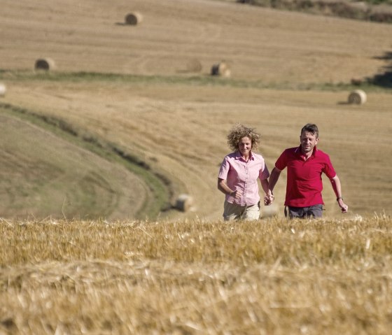 Ein Paar läuft Hand in Hand durch ein goldenes Feld. Im Hintergrund sind Heuballen auf den Feldern zu sehen., © Traumpfade/Kappest