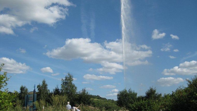 Ein Wasserfontäne sprüht hoch in den blauen Himmel. Der Himmel ist mit weißen Wolken bedeckt und im Vordergrund sind grüne Pflanzen zu sehen.