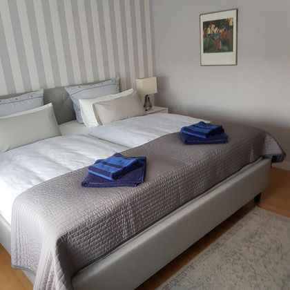 Ein modernes Schlafzimmer mit einem großen Doppelbett und eleganter Bettwäsche. Auf dem Bett liegen ordentliche Handtücher in Blau.