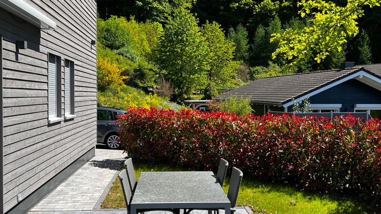 Ein schöne Terrasse mit einem Tisch und Stühlen. Im Hintergrund sind grüne Bäume und bunte Sträucher zu sehen.