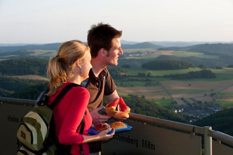 A couple are standing on a lookout tower and gazing into the distance. They are holding snacks in their hands. A hilly landscape can be seen in the background.