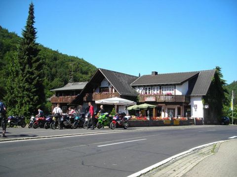 Ein gemütliches Gasthaus an einer Straße, umgeben von Bäumen. Motorräder stehen auf dem Parkplatz, während Menschen die Umgebung genießen.