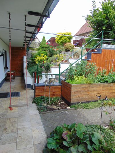 A well-kept garden with lush plants and vegetable beds. In the foreground, there is a swing and a staircase leading upwards.