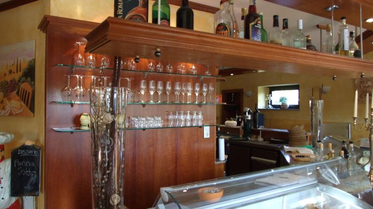 Eine gemütliche Bar mit einer Holztheke, Glasregalen und verschiedenen Getränken. Im Hintergrund sind Gläser und eine Kühlvitrine zu sehen.