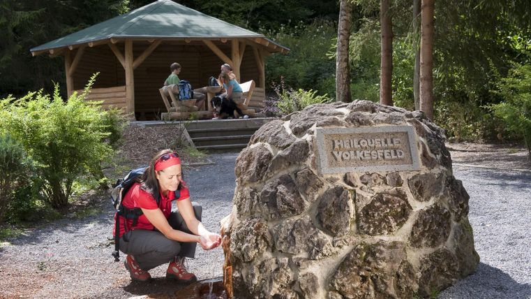 Eine Wandererin schöpft Wasser aus einer Felsquelle in der Natur. Im Hintergrund sitzen zwei Personen unter einem Pavillon und entspannen.