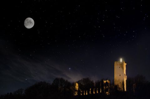 Eine beeindruckende Burgruine bei Nacht, beleuchtet von einem Vollmond. Der klare Himmel ist mit zahlreichen Sternen geschmückt.