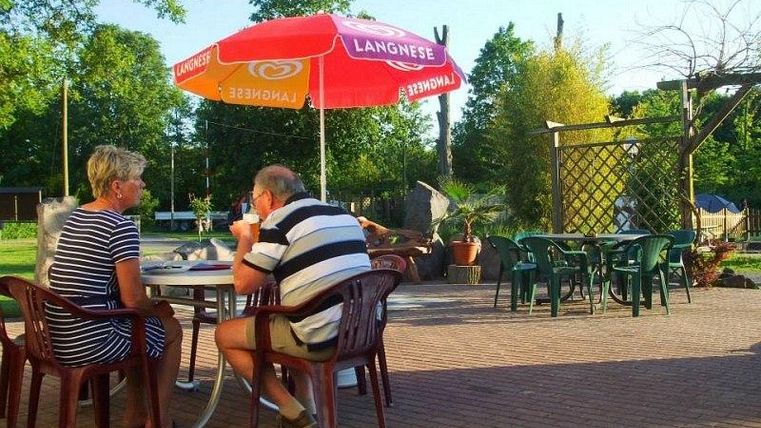 Ein Paar sitzt entspannt unter einem roten Sonnenschirm in einem Garten. Sie genießen Getränke an einem Tisch auf einem gut gepflegten Platz.