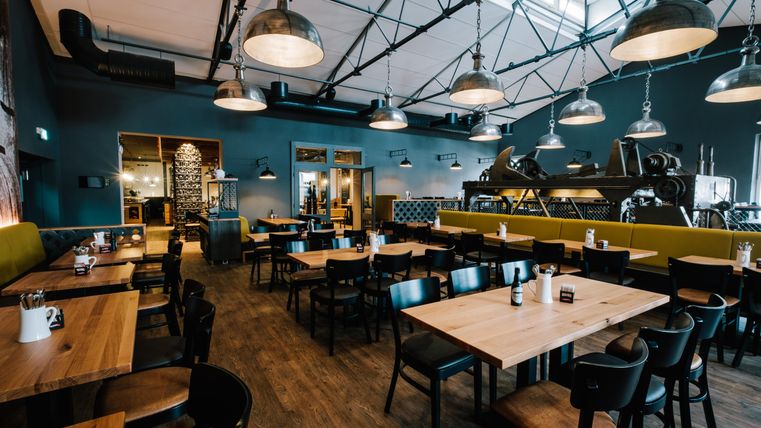 Ein modernes Restaurant mit Holztischen und schwarzen Stühlen. Es hat eine gemütliche Atmosphäre mit einer auffälligen Beleuchtung und einer offenen Küche.
