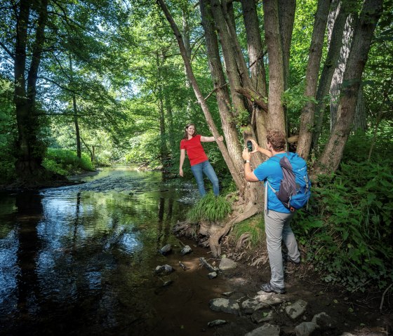Zwei Personen im Wald, eine posiert auf einem Baum, die andere fotografiert. Ein Bach flie&szlig;t im Hintergrund, umgeben von &uuml;ppigem Gr&uuml;n., &copy; kappest_remet