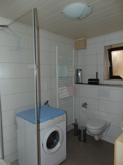 A modern bathroom with a shower, a washing machine, and a towel holder. The walls are tiled in a light color, and there is a window for natural light.