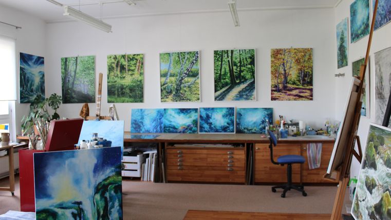 Ein Künstleratelier mit mehreren Malereien an den Wänden. Im Raum stehen Tische und ein Stuhl, um kreative Arbeiten durchzuführen.