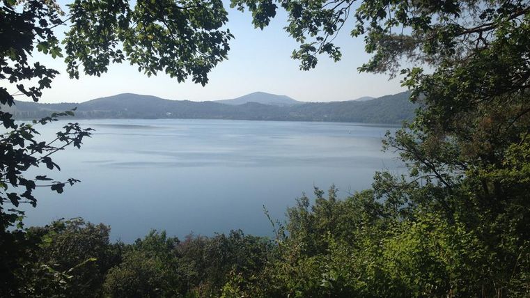 Ein ruhiger See, umgeben von Bäumen und Hügeln, unter einem klaren Himmel. Die Spiegelung des Himmels auf dem Wasser schafft eine friedliche Atmosphäre.
