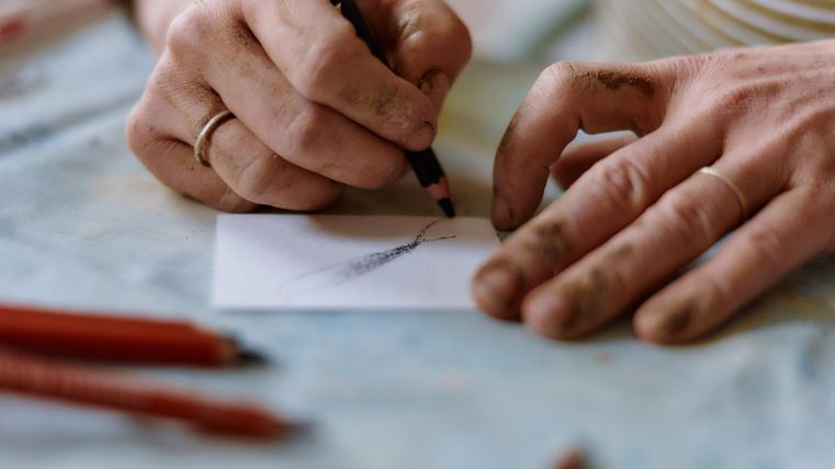 Eine Hand zeichnet mit einem Bleistift auf ein Blatt Papier. Rote Buntstifte sind im Hintergrund zu sehen.