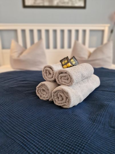Ein gemütliches Bett mit einem blauen Bettüberzug und mehreren Kissen. Darauf liegen sorgfältig aufgerollte Handtücher mit kleinen Dekorationen.