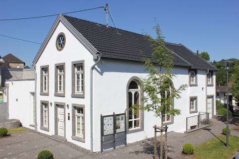 A bright, modern building with white walls and a dark roof. It is situated in a peaceful setting with small trees and a clear sky.