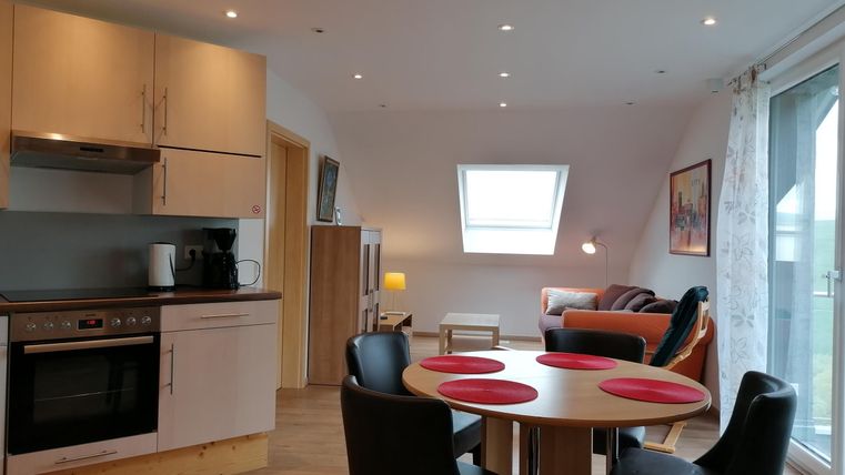 A modern kitchen with a dining table and four chairs. In the background, there is a cozy living area with a sofa and plenty of daylight.