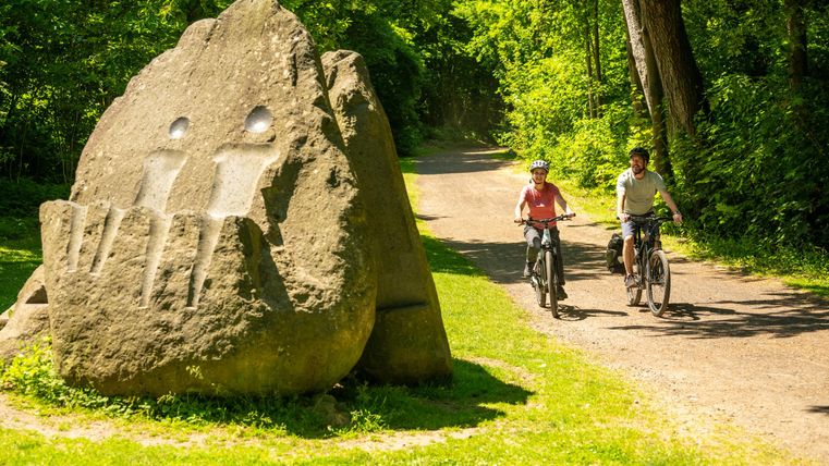 Zwei Radfahrer fahren auf einem Weg durch eine grüne Landschaft. Ein großer, markanter Stein mit einer besonderen Form steht daneben.