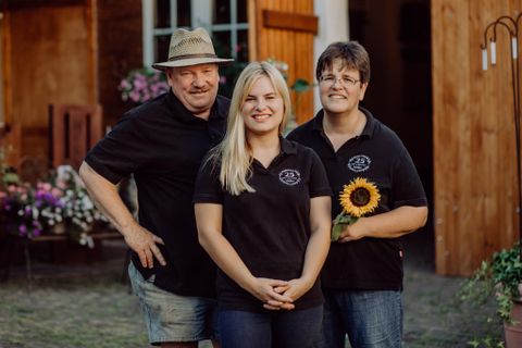 Three people stand in front of a wooden building. They are wearing black T-shirts and smiling, while one person is holding a sunflower in their hand.