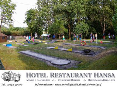 A mini golf course with various obstacles and many people playing outdoors. Surrounded by trees and a beautiful landscape.