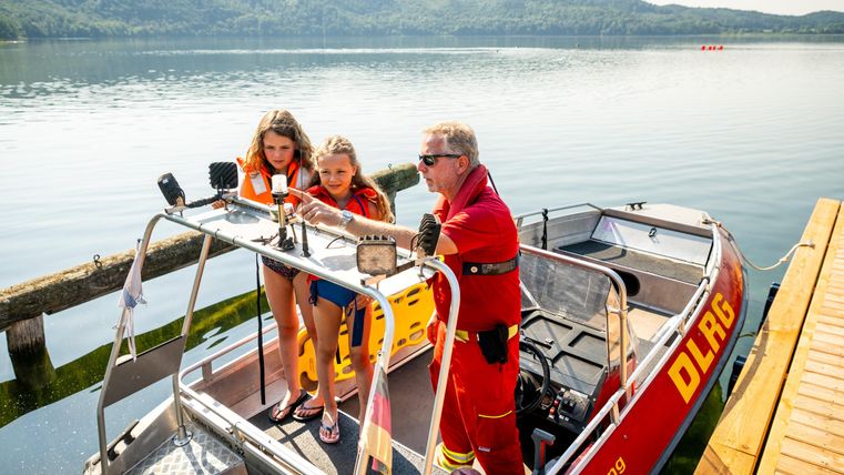 A rescue boat drill at the lake. Two children in life jackets stand next to a lifeguard on a boat.