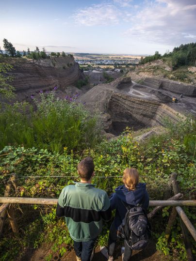 Two people are standing at a viewpoint, admiring an impressive landscape with layered rocks. In the background, buildings and a clear sky are visible.