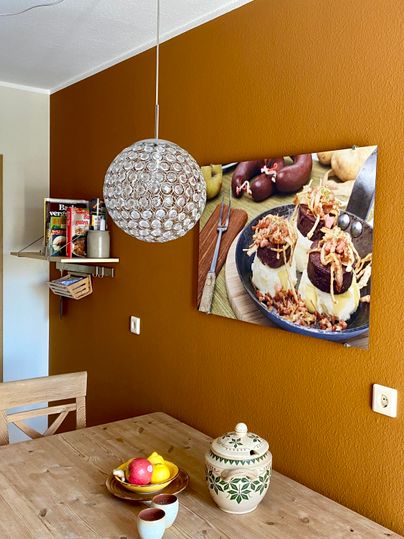 Eine gemütliche Küche mit einer orangenen Wandfarbe. Auf dem Tisch liegt eine Obstschale und an der Wand hängt ein Bild von köstlichen Desserts.