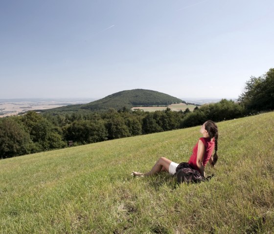 Eine Frau in rotem Shirt sitzt auf einer Wiese und schaut auf den bewaldeten Hochsimmer. Der Himmel ist klar und blau., © Traumpfade/Kappest