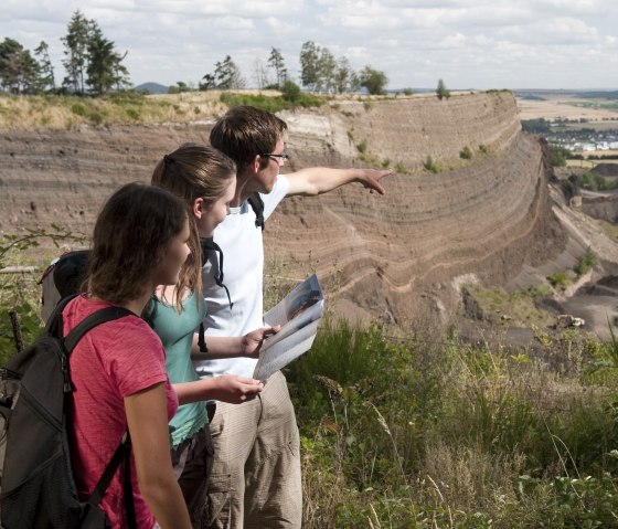 Drei Wanderer betrachten einen Steinbruch am Eppelsberg. Eine Person zeigt in die Ferne, während eine andere eine Karte hält. Im Hintergrund weite Landschaft., © Traumpfade/Kappest