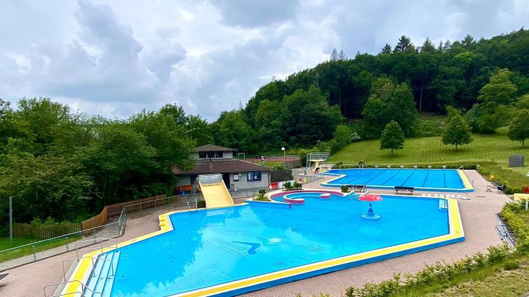 Ein Schwimmbad mit mehreren Becken und einer grünen Umgebung. Es gibt eine Liegewiese und eine gemütliche Atmosphäre unter einem bewölkten Himmel.