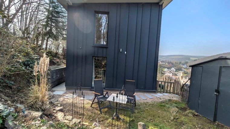 Ein modernes, schwarzes Haus auf einem Hügel umgeben von Bäumen. Im Vordergrund stehen zwei Stühle auf einer Terrasse mit Blick auf die Landschaft.
